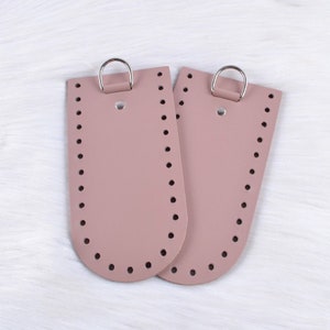 Op de afbeelding: Twee lederen tasbodem inzetstukken in een stoffige roze kleur met zilveren metalen ringen. De inzetstukken hebben een afgeronde bodem en zijn voorgeperforeerd met gaten voor het naaien.