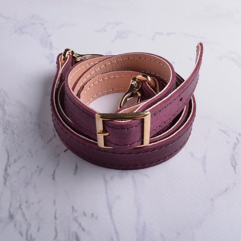 May include: A burgundy leather strap with a gold metal buckle and clasp. The strap is folded in a circle and is ready to be attached to a bag.