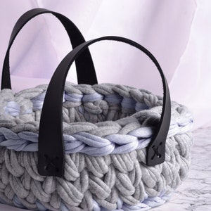 Peut inclure: Un panier en crochet gris et bleu clair avec des anses en cuir noir. Le panier est fait de fil épais et torsadé et a une large ouverture ronde.