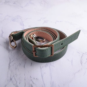 May include: A green leather strap with a brass buckle and two clasps. The strap is folded and coiled on a white surface.