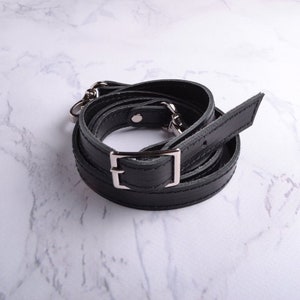 May include: Black leather strap with silver hardware and a rectangular buckle. The strap is coiled and ready to be attached to a bag.