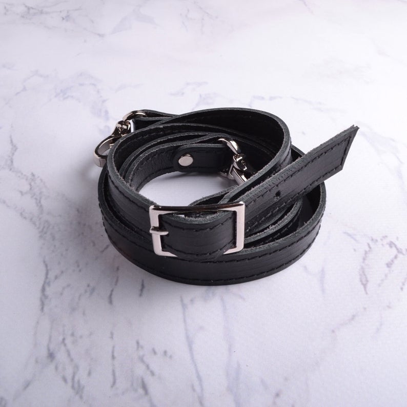 May include: Black leather strap with silver hardware and a rectangular buckle. The strap is coiled and ready to be attached to a bag.