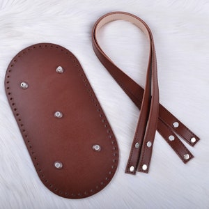 May include: A brown leather oval-shaped bag base with a stitched edge and metal rivets.  The base is accompanied by a brown leather strap with metal rivets.