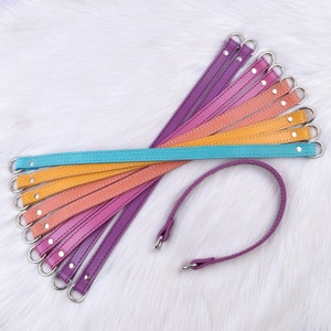 May include: A set of nine leather straps in various shades of purple, orange, and blue, with silver metal rings at each end. The straps are arranged in a rainbow pattern, with the lightest color at the top and the darkest color at the bottom. One strap is looped into a circle.