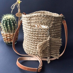 May include: A brown crocheted bag with a long brown leather strap. The bag has a fringe detail and a small pocket on the front.