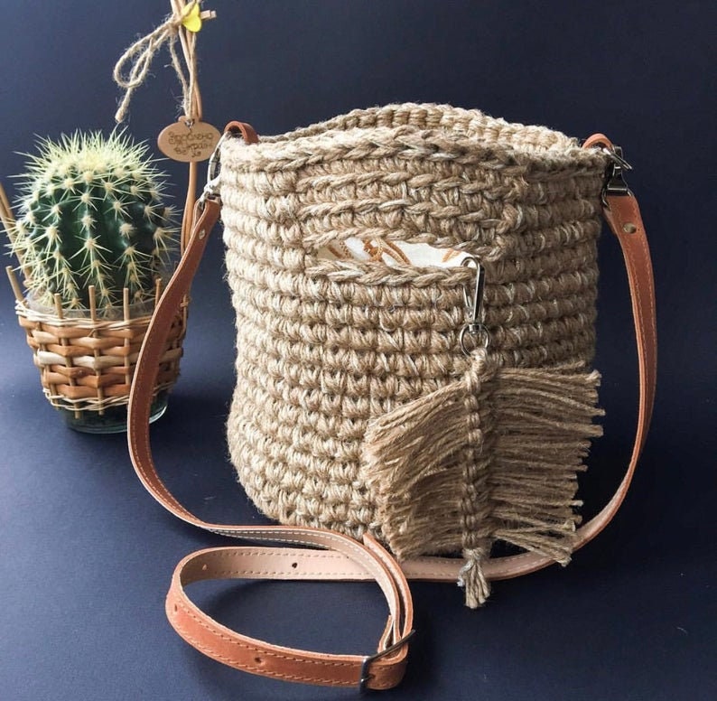 May include: A brown crocheted bag with a long brown leather strap. The bag has a fringe detail and a small pocket on the front.