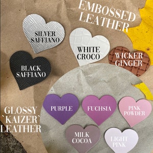 May include: A collection of heart-shaped leather samples in various colors and textures, including silver saffiano, black saffiano, white croco, wicker ginger, purple, fuchsia, pink powder, milk cocoa, and light pink. The samples are labeled with their names and grouped under the headings "Embossed Leather" and "Glossy Kaizer Leather".