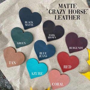 May include: Nine heart-shaped leather swatches in various colors, including black, green, tan, blue, azure, coral, red, burgundy, and dark brown. The swatches are arranged in a circle on a brown paper background. The text "MATTE 'CRAZY HORSE' LEATHER" is written in black at the top of the image.