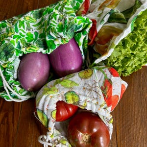 May include: Three reusable produce bags filled with fresh vegetables. Two purple eggplants are in a green leaf-patterned bag. Red tomatoes are in a bag with a tomato and lime print. Green lettuce is visible.