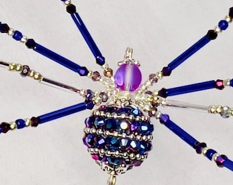 May include: A decorative beaded spider ornament. The spider's body is made of iridescent blue and purple beads, with dark blue legs and a purple bead head. The legs are made of dark blue and silver beads. A unique, handcrafted item.
