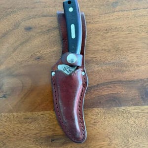 Könnte beinhalten: Ein schwarzes und silbernes Messer mit einem dunkelbraunen Griff steckt in einer dunkelbraunen Lederscheide. Die Scheide hat einen genähten Rand und einen Metallverschluss. Das Messer und die Scheide liegen auf einer Holzoberfläche.