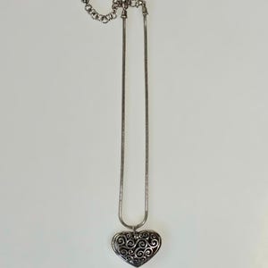 May include: A silver-toned necklace featuring a snake chain and a heart-shaped pendant. The pendant displays an intricate, openwork filigree pattern with dark recessed areas. The chain includes an adjustable extender with a lobster claw clasp.