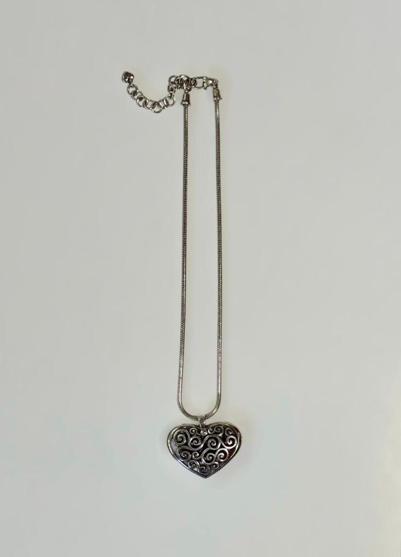 May include: A silver-toned necklace featuring a snake chain and a heart-shaped pendant. The pendant displays an intricate, openwork filigree pattern with dark recessed areas. The chain includes an adjustable extender with a lobster claw clasp.