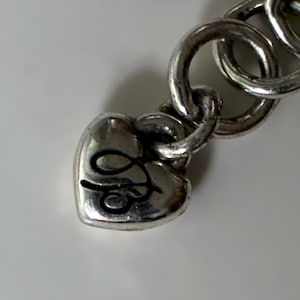 May include: A silver-toned metal bracelet featuring a heart-shaped charm attached to a series of circular links. The heart charm displays a black engraved cursive letter design on its surface, with a polished finish throughout the metal components.