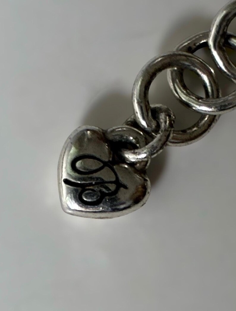 May include: A silver-toned metal bracelet featuring a heart-shaped charm attached to a series of circular links. The heart charm displays a black engraved cursive letter design on its surface, with a polished finish throughout the metal components.