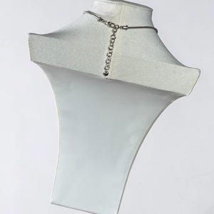 May include: A silver-toned necklace chain featuring a snake chain design and a lobster claw clasp. An adjustable extender chain is attached to the clasp, ending with a small heart-shaped charm. The metal has a polished, reflective finish.