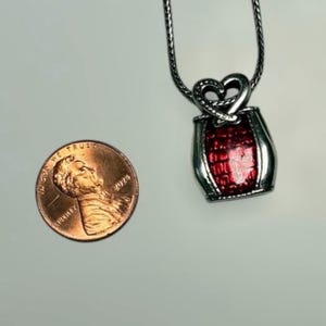 May include: A silver-toned pendant necklace featuring a heart-shaped top and a rectangular base with a textured, glossy red inlay. The pendant hangs from a thin silver chain and is shown next to a 2024 United States one-cent copper coin.