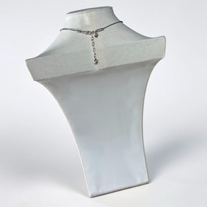 May include: A silver-toned necklace featuring a thin chain and a central decorative element with a dangling extension chain. The necklace is displayed on a white fabric-covered bust, showcasing the back closure mechanism and the adjustable length.