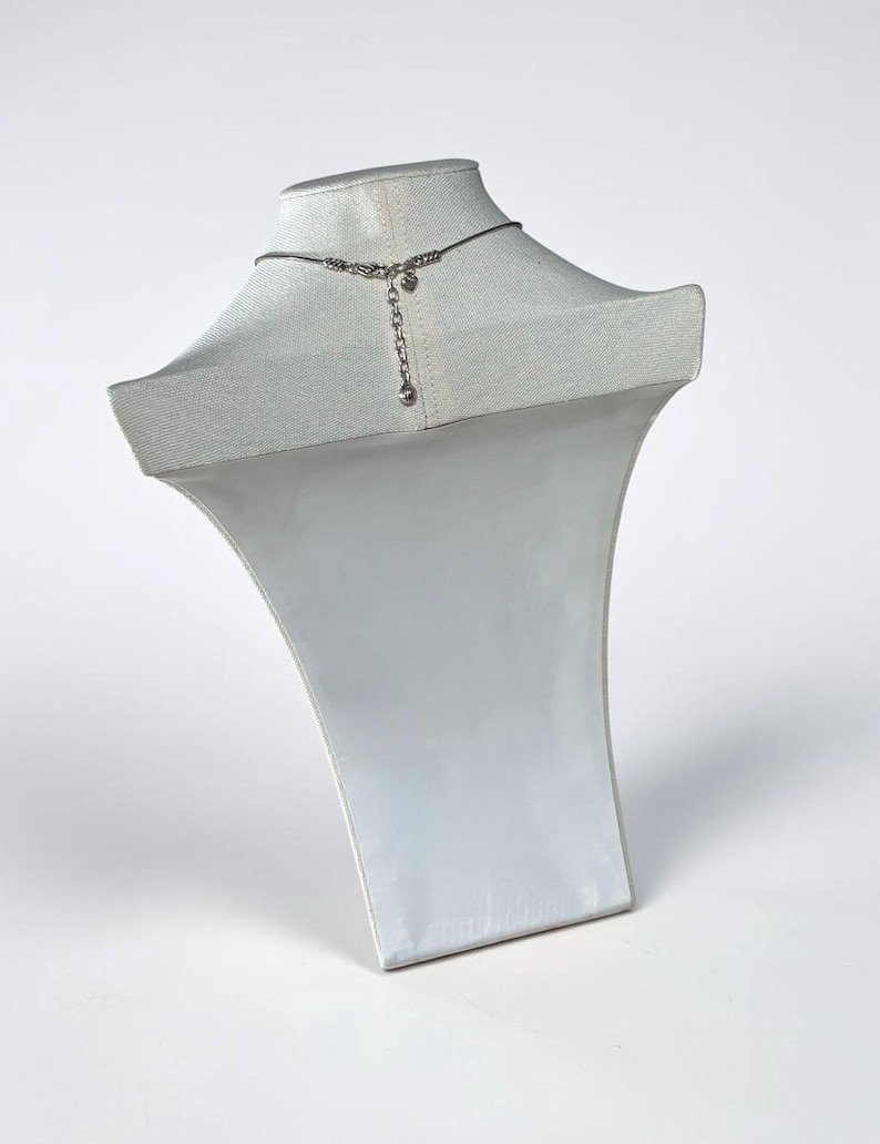 May include: A silver-toned necklace featuring a thin chain and a central decorative element with a dangling extension chain. The necklace is displayed on a white fabric-covered bust, showcasing the back closure mechanism and the adjustable length.