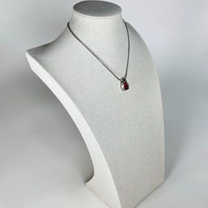 May include: A silver-toned chain necklace featuring a small, teardrop-shaped pendant. The pendant has a polished metallic frame surrounding a vibrant, deep red center stone. The necklace rests on a neutral-colored, textured display bust.