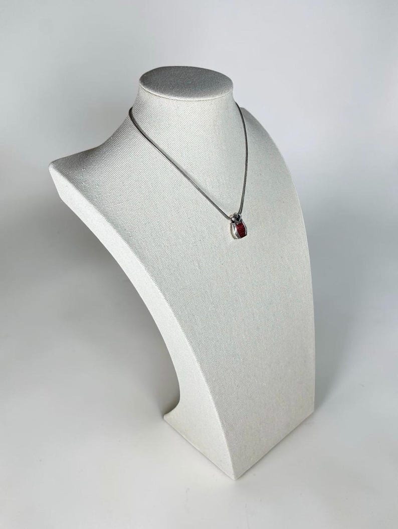 May include: A silver-toned chain necklace featuring a small, teardrop-shaped pendant. The pendant has a polished metallic frame surrounding a vibrant, deep red center stone. The necklace rests on a neutral-colored, textured display bust.