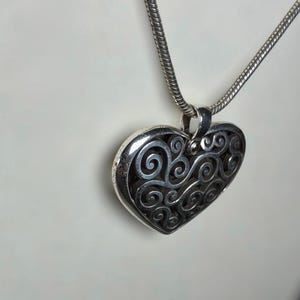 May include: A silver-toned heart-shaped pendant featuring an intricate openwork filigree design with swirling patterns. The pendant is suspended from a matching silver-toned snake chain necklace, showcasing a polished metallic finish throughout.