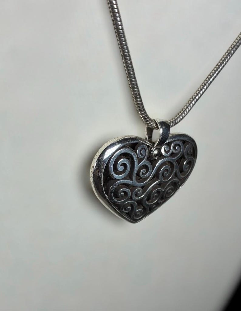 May include: A silver-toned heart-shaped pendant featuring an intricate openwork filigree design with swirling patterns. The pendant is suspended from a matching silver-toned snake chain necklace, showcasing a polished metallic finish throughout.