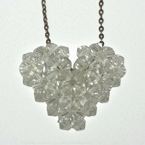 May include: A clear crystal heart pendant necklace. The heart is made of many small, faceted crystals clustered together. The necklace chain is a silver color and attaches to the top of the heart.