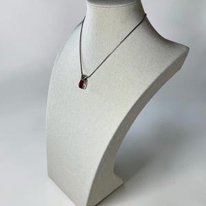 May include: A silver-toned necklace featuring a small, teardrop-shaped pendant with a deep red stone inlay. The pendant is suspended from a thin, delicate silver-toned chain that rests against the neck form, showcasing a polished metallic finish.
