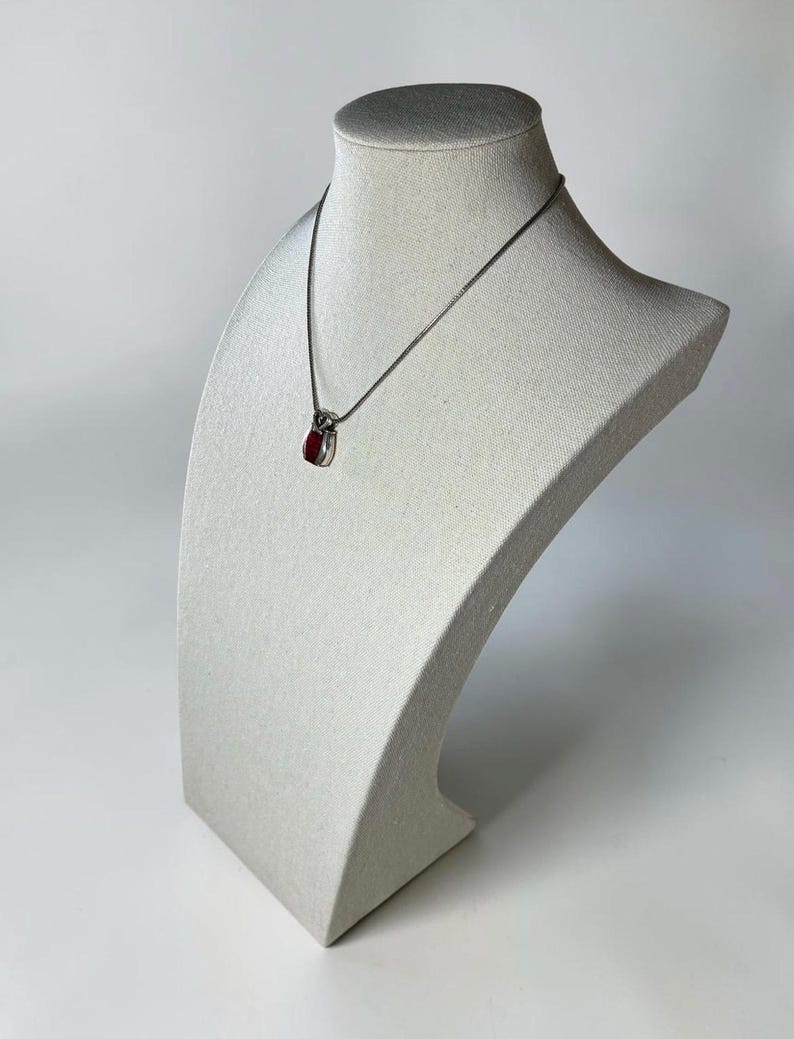 May include: A silver-toned necklace featuring a small, teardrop-shaped pendant with a deep red stone inlay. The pendant is suspended from a thin, delicate silver-toned chain that rests against the neck form, showcasing a polished metallic finish.