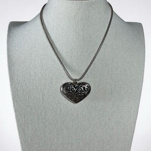 May include: A silver-toned necklace featuring a heart-shaped pendant with an intricate, dark-oxidized filigree scrollwork pattern. The pendant hangs from a smooth, round snake chain, creating a cohesive metallic piece with a polished finish.