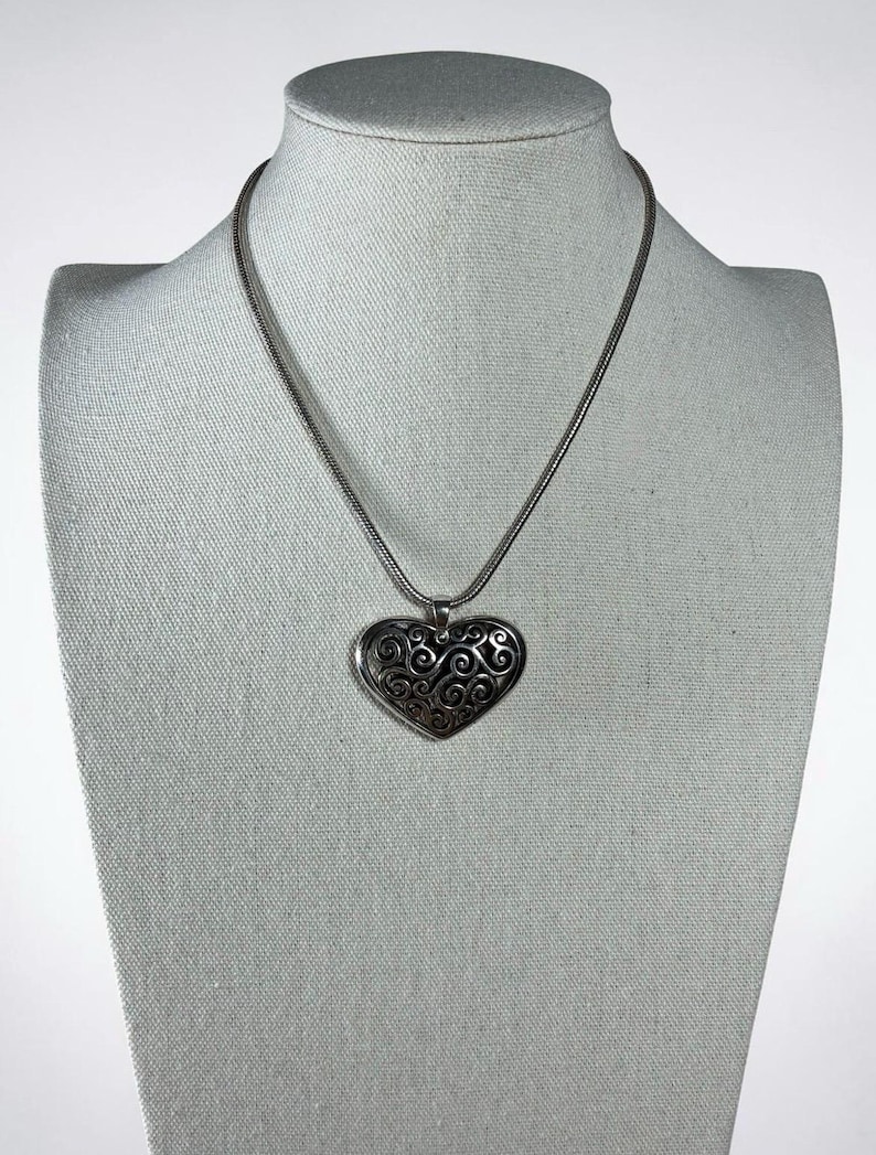 May include: A silver-toned necklace featuring a heart-shaped pendant with an intricate, dark-oxidized filigree scrollwork pattern. The pendant hangs from a smooth, round snake chain, creating a cohesive metallic piece with a polished finish.