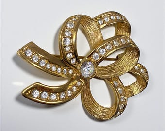 May include: A gold-toned bow brooch with a textured surface, adorned with clear, sparkling rhinestones. The brooch features a central, larger rhinestone and is designed to be worn as a decorative accessory.