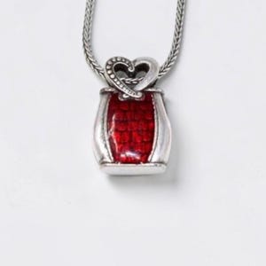 May include: A silver-toned pendant necklace featuring a rectangular base with a vibrant red, textured inlay. The top of the pendant is shaped like a stylized heart with a dotted pattern, suspended from a matching silver-toned wheat chain.