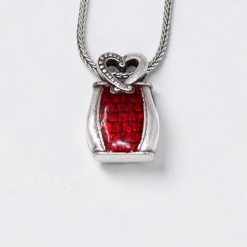 May include: A silver-toned pendant necklace featuring a rectangular base with a vibrant red, textured inlay. The top of the pendant is shaped like a stylized heart with a dotted pattern, suspended from a matching silver-toned wheat chain.