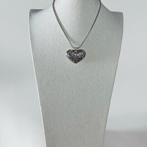May include: A silver-toned heart-shaped pendant necklace featuring an intricate, dark-oxidized filigree swirl pattern. The pendant hangs from a smooth, round silver-toned chain, creating a cohesive metallic look with a polished finish and detailed texture.