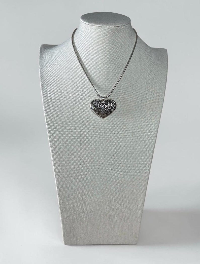 May include: A silver-toned heart-shaped pendant necklace featuring an intricate, dark-oxidized filigree swirl pattern. The pendant hangs from a smooth, round silver-toned chain, creating a cohesive metallic look with a polished finish and detailed texture.