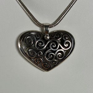 May include: A silver-toned heart-shaped pendant featuring an intricate openwork design of swirling, interconnected metal scrolls. The pendant is suspended from a matching silver-toned snake chain necklace that loops through a polished metal bail.