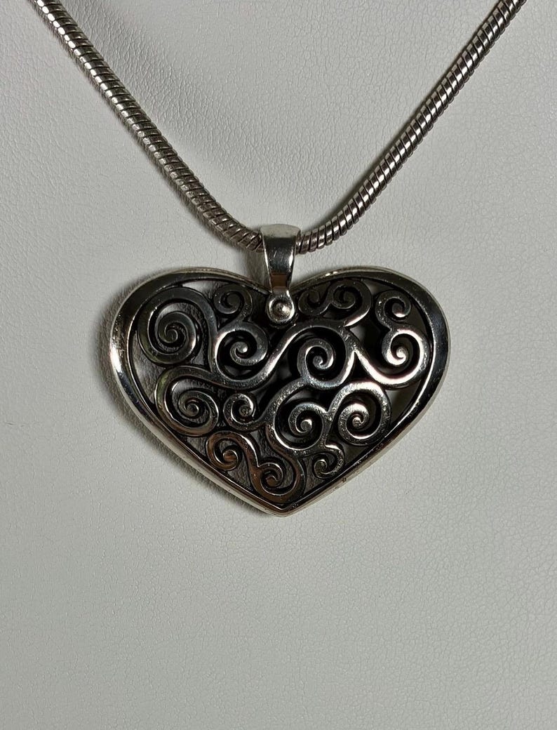 May include: A silver-toned heart-shaped pendant featuring an intricate openwork design of swirling, interconnected metal scrolls. The pendant is suspended from a matching silver-toned snake chain necklace that loops through a polished metal bail.
