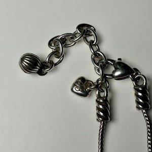 May include: A silver-toned necklace featuring a chain with a lobster claw clasp, a small heart-shaped charm, and a larger ribbed spherical bead at the end of an extender chain. The metal components have a polished finish with visible textured detailing.