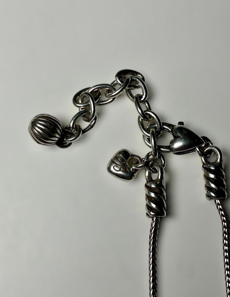 May include: A silver-toned necklace featuring a chain with a lobster claw clasp, a small heart-shaped charm, and a larger ribbed spherical bead at the end of an extender chain. The metal components have a polished finish with visible textured detailing.