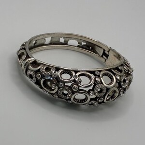 May include: Silver-toned hinged bangle bracelet with an openwork design of circles and ovals. The bracelet is embellished with small, clear crystals. The clasp is visible on the side. This jewelry piece is suitable for everyday wear or special occasions.