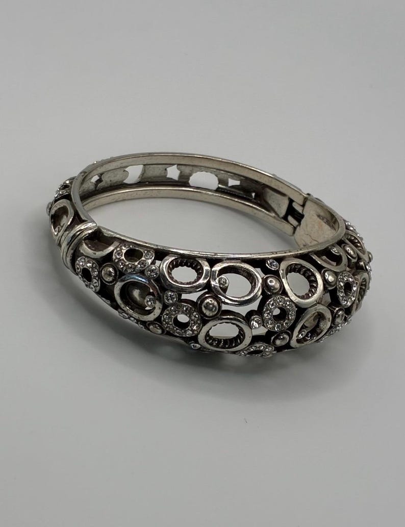 May include: Silver-toned hinged bangle bracelet with an openwork design of circles and ovals. The bracelet is embellished with small, clear crystals. The clasp is visible on the side. This jewelry piece is suitable for everyday wear or special occasions.