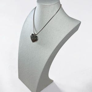 May include: A silver-toned necklace featuring a heart-shaped pendant with an intricate, dark-toned filigree scrollwork pattern. The pendant hangs from a thin, smooth silver-toned chain, creating a classic and detailed piece of jewelry.