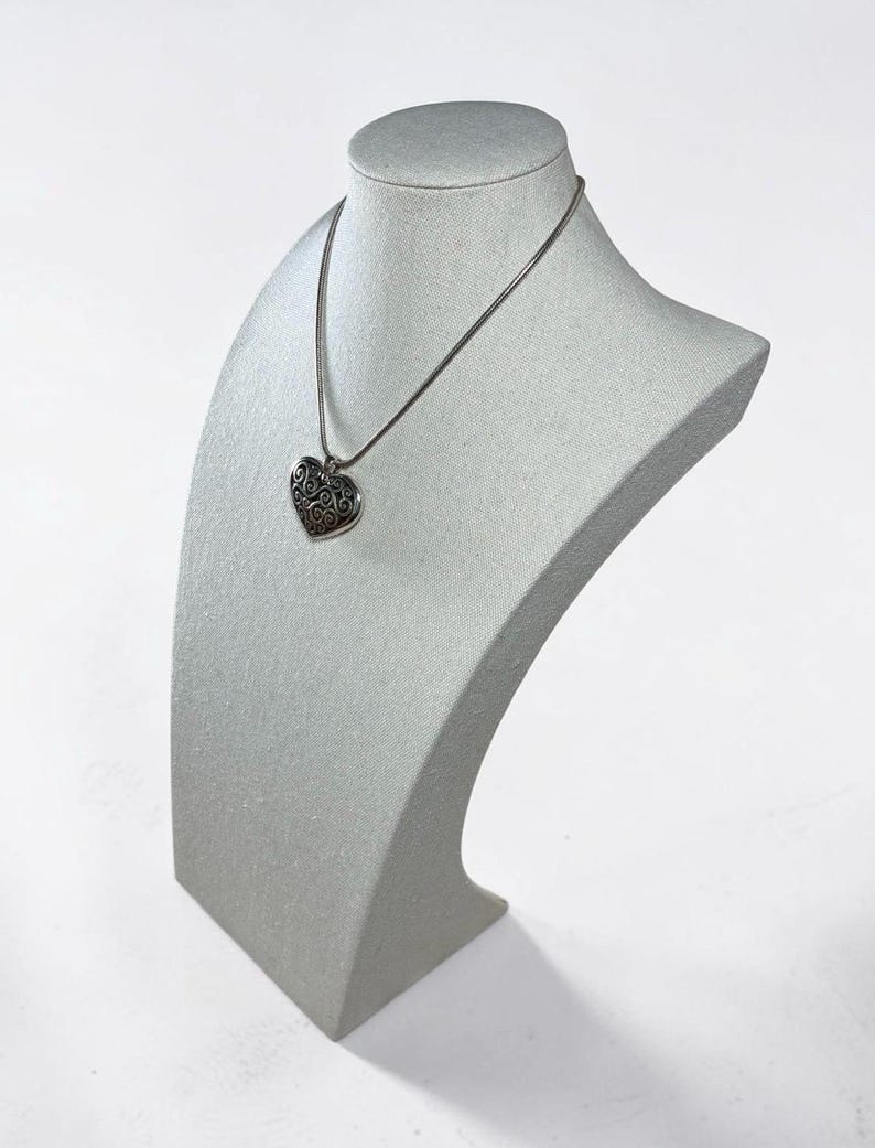 May include: A silver-toned necklace featuring a heart-shaped pendant with an intricate, dark-toned filigree scrollwork pattern. The pendant hangs from a thin, smooth silver-toned chain, creating a classic and detailed piece of jewelry.