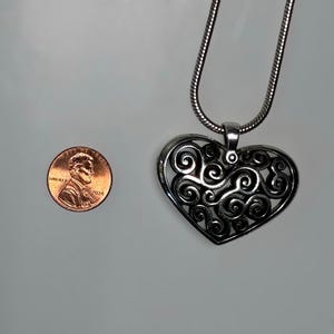 May include: A silver-toned heart-shaped pendant featuring an intricate openwork filigree design of swirling patterns, suspended from a matching snake chain necklace, shown next to a 2024 United States copper-colored penny for scale.
