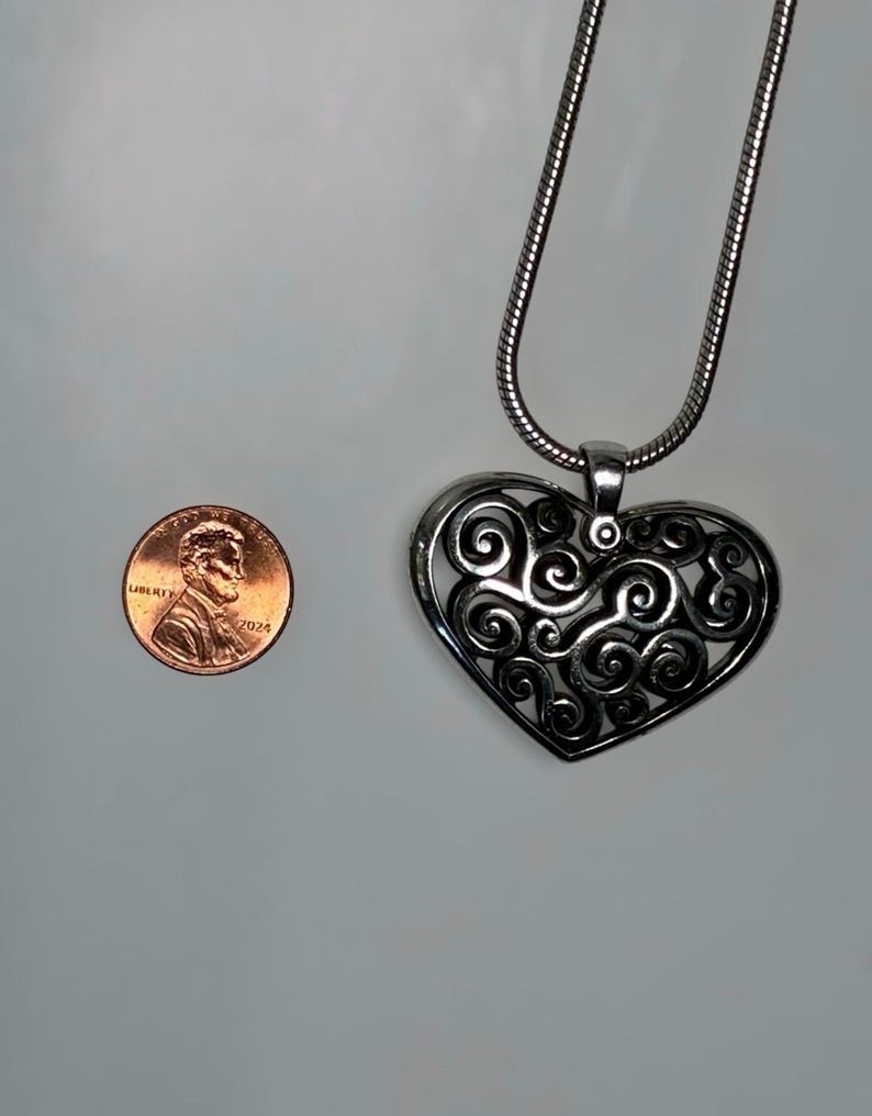 May include: A silver-toned heart-shaped pendant featuring an intricate openwork filigree design of swirling patterns, suspended from a matching snake chain necklace, shown next to a 2024 United States copper-colored penny for scale.