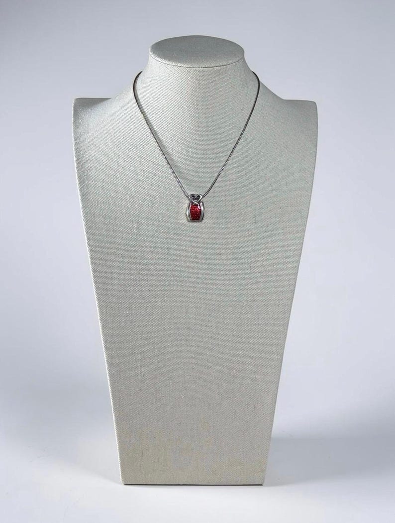 May include: A silver-toned necklace featuring a small, rectangular pendant with a vibrant red center inlay. The pendant is suspended from a thin, delicate silver-toned chain, creating a simple and refined piece of jewelry for everyday wear.