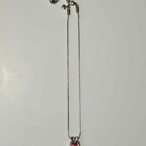 May include: A silver-toned snake chain necklace featuring a small, rectangular pendant with a textured, bright red center and a heart-shaped top. The necklace includes an adjustable chain with a small dangling charm at the clasp end.