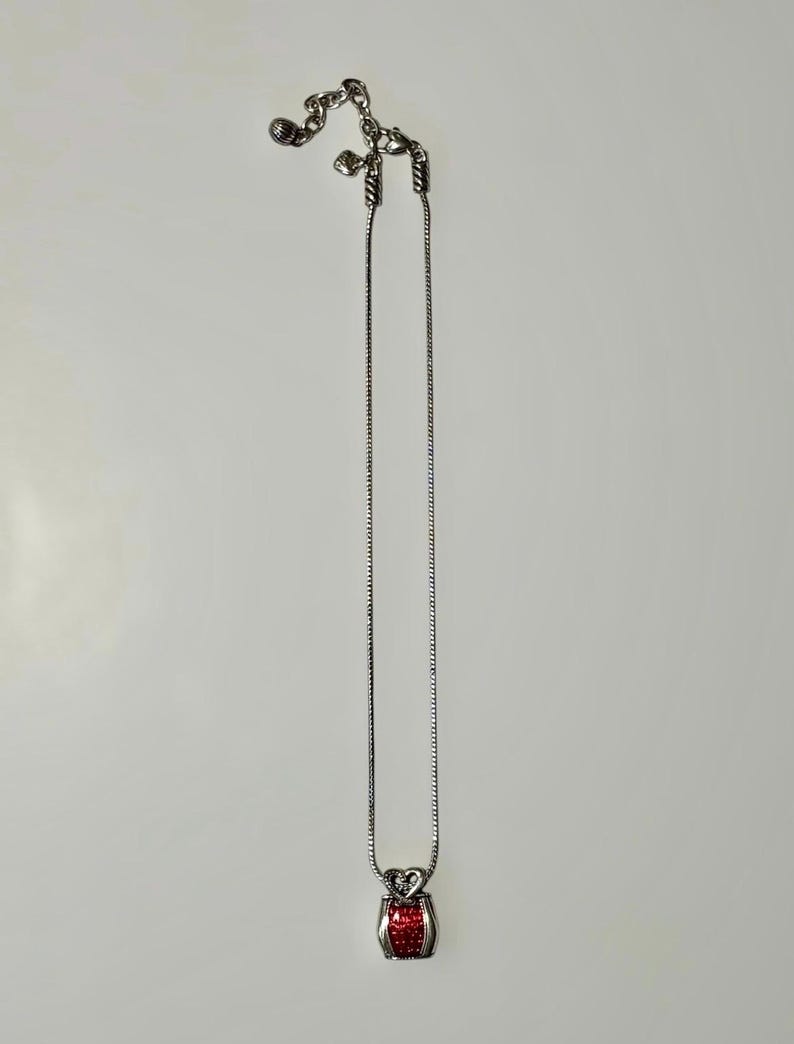 May include: A silver-toned snake chain necklace featuring a small, rectangular pendant with a textured, bright red center and a heart-shaped top. The necklace includes an adjustable chain with a small dangling charm at the clasp end.